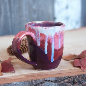May include: A handmade ceramic mug with a unique glaze. The mug has a deep purple base that transitions to shades of red and white with blue drips. The handle is a gradient of red and purple.