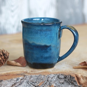 Peut inclure: Une tasse en céramique avec un vernis brillant dans des tons bleu marine profond, bleu roi et turquoise plus clair. La tasse présente un corps cylindrique avec une anse incurvée sur le côté droit et un bord lisse et arrondi, aux nuances variées.
