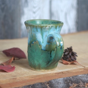 May include: A ceramic mug featuring a reactive glaze with a blend of deep forest green at the base transitioning into a lighter sky blue and white near the rim. The mug has a sturdy handle and a slightly flared opening with a smooth, glossy finish.