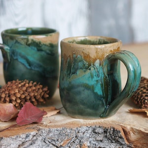 May include: Two ceramic mugs featuring a reactive glaze with blended shades of deep forest green, teal, and a warm tan rim. Each mug has a sturdy, curved handle and a rounded body, showcasing a smooth, glossy finish with organic colour transitions.