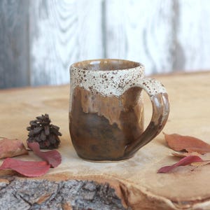 May include: A ceramic mug featuring a two-tone glaze with a dark brown base that transitions into a lighter, speckled cream-coloured rim. The mug has a rounded body and a curved handle, showcasing a smooth, glossy finish on the exterior surface.
