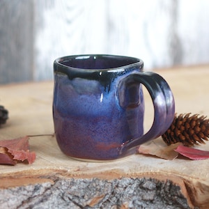 Peut inclure: Une tasse en céramique avec un corps arrondi et une anse incurvée. L'extérieur présente un vernis brillant qui passe d'un violet profond à la base à un bleu vif au centre, surmonté d'un bord noir foncé qui coule sur les côtés.