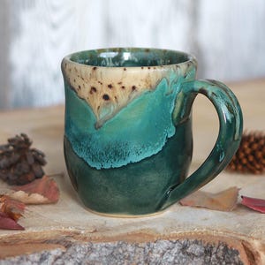 May include: A ceramic mug featuring a reactive glaze with a cream-coloured rim that transitions into vibrant turquoise and deep forest green tones. The mug has a rounded body and a curved handle, showcasing a smooth, glossy finish throughout the exterior.