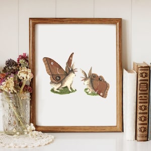 May include: A framed print of two brown rabbits with butterfly wings. The rabbits are facing each other and are on a white background. The frame is brown wood.