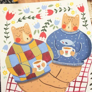 May include: An illustration on paper featuring two bears sitting together. One bear wears a multicolored patchwork sweater, the other a blue sweater with a fish graphic. Both hold white mugs with red tulip designs, surrounded by red and blue flowers.