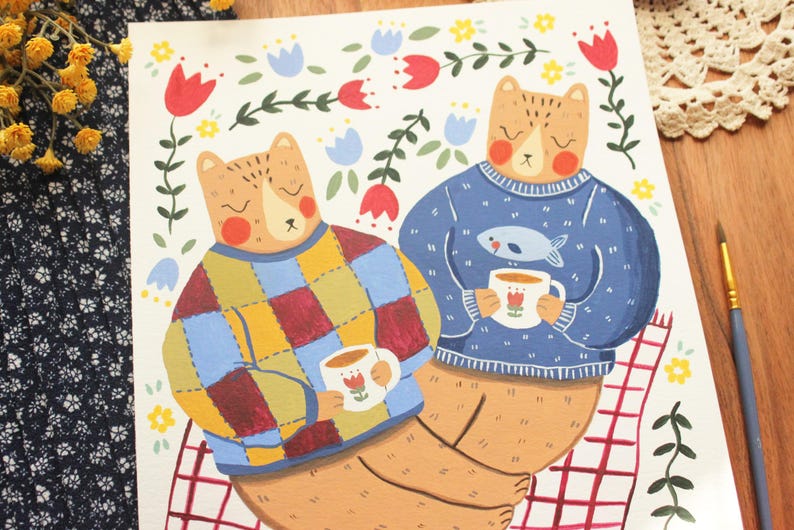 May include: An illustration on paper featuring two bears sitting together. One bear wears a multicolored patchwork sweater, the other a blue sweater with a fish graphic. Both hold white mugs with red tulip designs, surrounded by red and blue flowers.