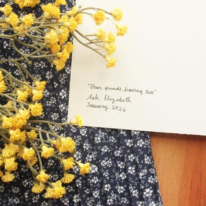 May include: A white card with the handwritten text "Bear friends having tea", "Ash Elizabeth", and "January 2026" in black ink. The card is placed next to a cluster of small yellow flowers and a dark blue fabric with a white floral pattern.