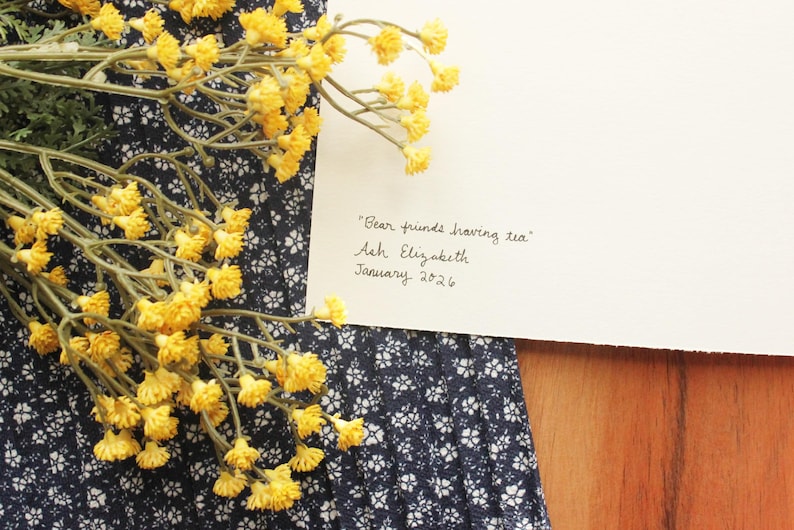 May include: A white card with the handwritten text "Bear friends having tea", "Ash Elizabeth", and "January 2026" in black ink. The card is placed next to a cluster of small yellow flowers and a dark blue fabric with a white floral pattern.