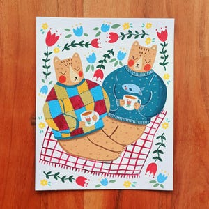 May include: A rectangular art print featuring two tan bears sitting on a red and white checkered blanket. One bear wears a multicolored checkered sweater, the other a teal sweater with a fish graphic. Both hold white mugs with tulip designs.