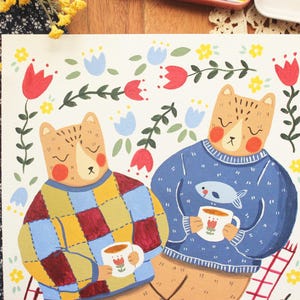 May include: A square art print featuring two bears sitting together. One bear wears a multicolored checkered sweater, the other a blue sweater with a fish graphic. Both hold white mugs with tulip designs, surrounded by red and blue floral motifs.