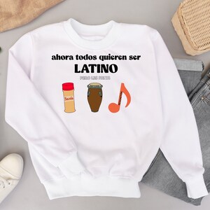 Könnte beinhalten: Weißes Sweatshirt mit dem Text "ahora todos quieren ser LATINO pero les falta". Das Design umfasst Illustrationen eines Gewürzbehälters, einer Trommel und einer Musiknote mit dem Wort "Reggaeton".