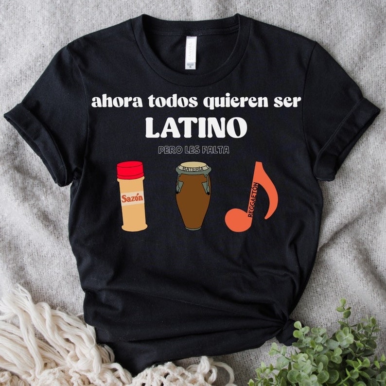 Puede incluir: Camiseta negra con el texto "ahora todos quieren ser LATINO pero les falta." La camiseta presenta ilustraciones de un bote de especias, un tambor y una nota musical con la palabra "Reggaeton."