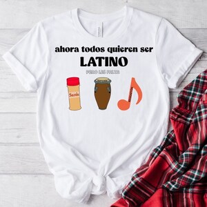 Puede incluir: Camiseta blanca con el texto "ahora todos quieren ser LATINO pero les falta". La camiseta presenta ilustraciones de un bote de especias, un tambor y una nota musical con la palabra "Reggaeton".