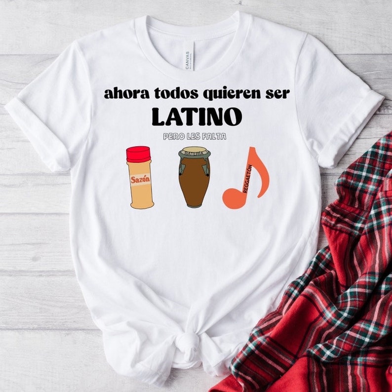 Puede incluir: Camiseta blanca con el texto "ahora todos quieren ser LATINO pero les falta". La camiseta presenta ilustraciones de un bote de especias, un tambor y una nota musical con la palabra "Reggaeton".