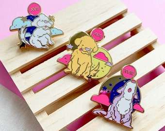 Puede incluir: Tres pines de esmalte con perros de dibujos animados en diferentes poses. Cada pin tiene un círculo rosa con números romanos en la parte superior de una luna azul, amarilla o morada. Los pines están sobre una superficie de madera.