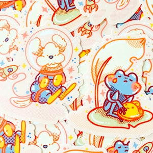 May include: A set of colourful stickers featuring a white dog wearing a blue and orange spacesuit, a blue frog sitting on a green lily pad, and a rocket ship. The stickers are on a white background with pink, yellow, and blue accents.