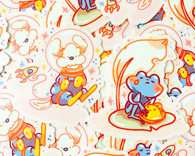 May include: A set of colourful stickers featuring a white dog wearing a blue and orange spacesuit, a blue frog sitting on a green lily pad, and a rocket ship. The stickers are on a white background with pink, yellow, and blue accents.