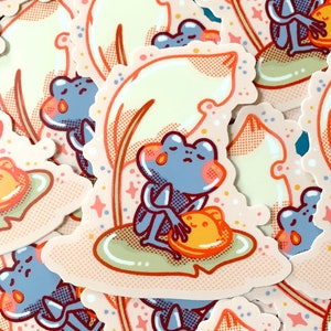May include: A sticker featuring a blue frog sitting on a lily pad with a yellow mushroom. The frog is holding the mushroom in its hands. The sticker has a light pink background with a yellow leaf and white stars.