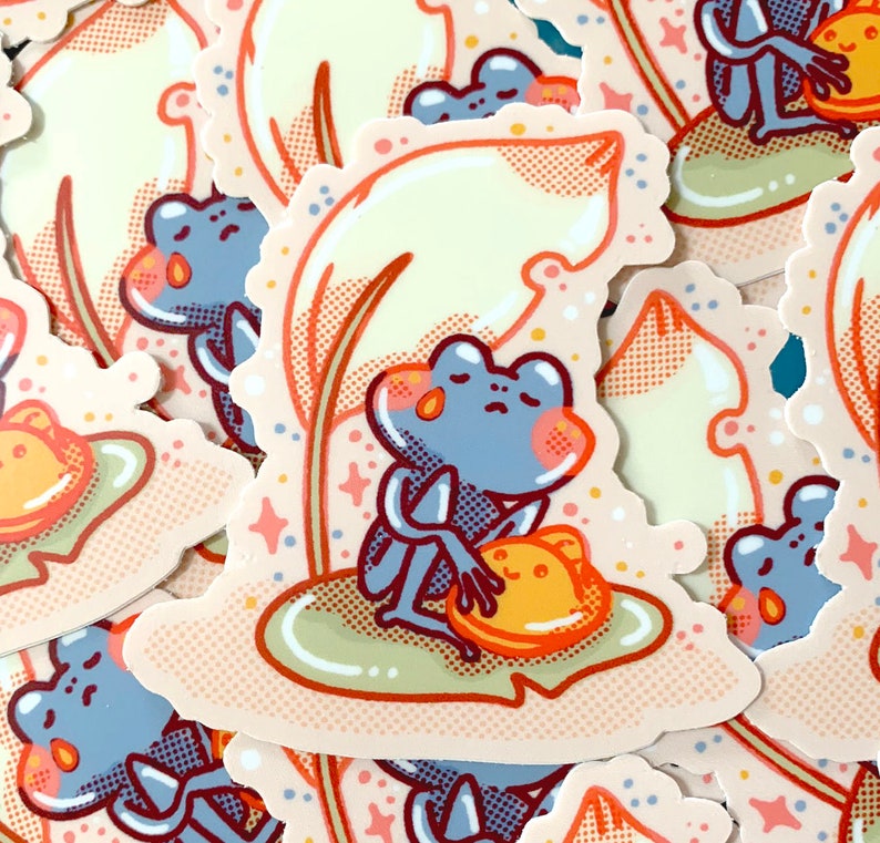 May include: A sticker featuring a blue frog sitting on a lily pad with a yellow mushroom. The frog is holding the mushroom in its hands. The sticker has a light pink background with a yellow leaf and white stars.