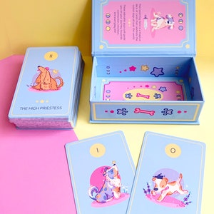Puede incluir: Una baraja de tarot Good Dog con una caja azul y cartas con ilustraciones de perros. La caja est&aacute; abierta y las cartas est&aacute;n dispuestas en forma de abanico. Las cartas est&aacute;n numeradas y tienen nombres como "La Suma Sacerdotisa" y "El Loco".