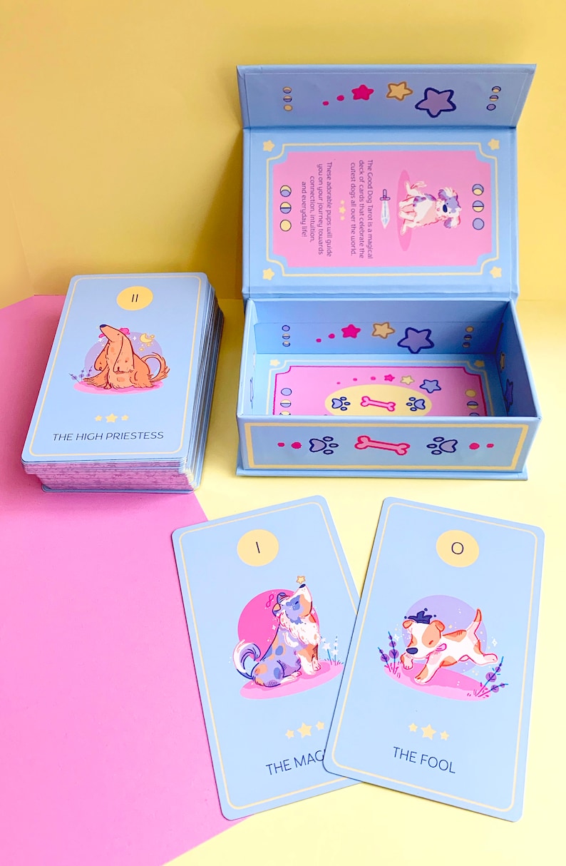 Puede incluir: Una baraja de tarot Good Dog con una caja azul y cartas con ilustraciones de perros. La caja est&aacute; abierta y las cartas est&aacute;n dispuestas en forma de abanico. Las cartas est&aacute;n numeradas y tienen nombres como "La Suma Sacerdotisa" y "El Loco".