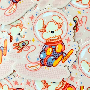 May include: A sticker featuring a cartoon dog wearing a space suit and helmet, floating in space with a rocket and stars. The sticker has a pink and white background with a dotted pattern.