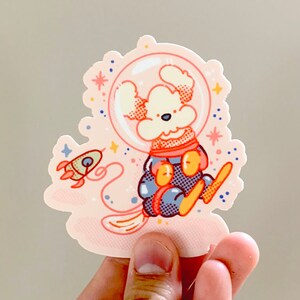 May include: A white dog wearing a blue and orange astronaut suit with a helmet and a rocket ship in the background. The sticker is on a pink background with white stars.