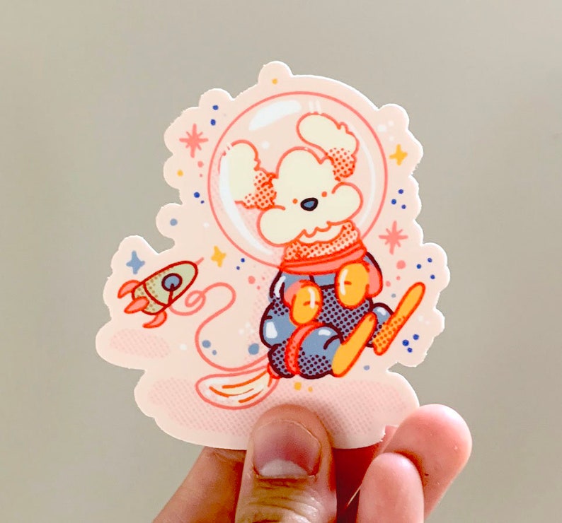 May include: A white dog wearing a blue and orange astronaut suit with a helmet and a rocket ship in the background. The sticker is on a pink background with white stars.