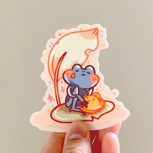 May include: A blue frog with a tear on its cheek sits on a green lily pad with a yellow and orange flower. The frog is holding the flower in its arms. The lily pad is surrounded by a pink and white background with white stars.