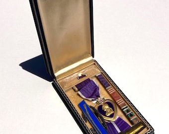 May include: A Purple Heart military medal set presented in a black rectangular display case with a cream-colored interior. The set includes a heart-shaped gold-tone medal with a purple enamel center, a purple ribbon, a blue bar, and a striped ribbon.