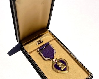 Peut inclure: Une médaille militaire Purple Heart présentant un profil doré de George Washington sur un fond violet en forme de cœur, suspendue à un ruban violet, présentée dans un coffret rectangulaire bleu foncé avec une doublure intérieure crème.