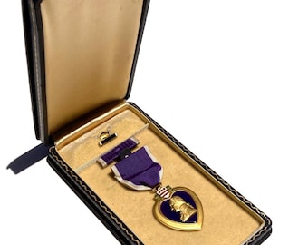 May include: A Purple Heart military medal displayed inside a dark blue rectangular presentation case with a cream-colored interior. The medal features a gold-toned heart-shaped frame with a purple enamel center, a profile portrait, and a purple ribbon.