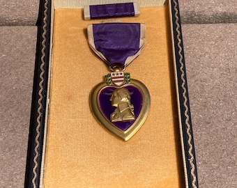 May include: A Purple Heart medal in a presentation case. The heart-shaped medal features a bust of George Washington and is suspended from a purple ribbon. The case is lined with gold fabric and has a black border.