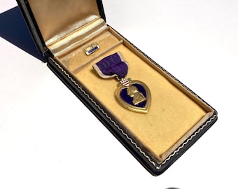 May include: A Purple Heart military medal featuring a gold profile of George Washington on a purple enamel heart, suspended from a purple ribbon with white edges, housed in a black rectangular presentation case with a tan velvet interior lining.