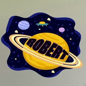 Peut inclure: Un autocollant mural sur le thème de l'espace bleu et jaune avec le nom "ROBERT" en texte noir sur une planète jaune avec des anneaux.