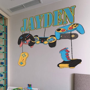May include: A child's bedroom with a climbing structure, a small table, and two chairs. The wall features a decorative design with the name "JAYDEN" and cartoon game controllers in blue, black, and yellow.