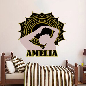 Könnte beinhalten: Ein Wandtattoo mit einer Yoga-Pose-Silhouette in einem schwarzen und goldenen Mandala-Design. Der Name "AMELIA" wird in fetten, schwarzen Buchstaben angezeigt. Das Tattoo befindet sich über einem Bett mit gestreifter Bettwäsche.