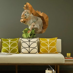 May include: A geometric design of a squirrel in shades of brown, gray, and green, sitting on a green leaf. The squirrel is on a wall behind a white couch with three pillows. The pillows have a green and white floral pattern.
