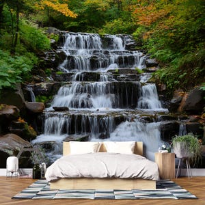May include: A bedroom with a waterfall mural. The mural depicts a multi-tiered waterfall cascading over dark rocks, surrounded by lush green and yellow foliage. A bed with white bedding and a wooden frame is in front of the mural.