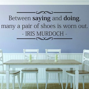May include: A dining room scene with a light blue wall featuring a quote by Iris Murdoch: "Between saying and doing, many a pair of shoes is worn out." A wooden table and four white chairs are in front.