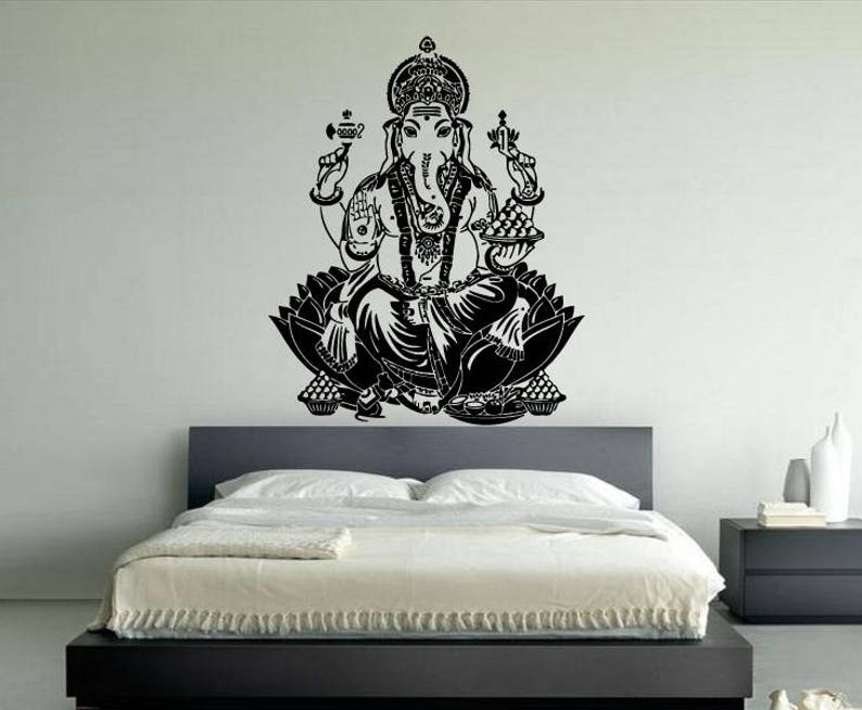May include: Black and white wall decal of Ganesha, the Hindu god of wisdom and prosperity, sitting in a lotus position. The decal is on a white wall above a black bed frame.