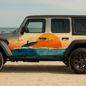 Op de afbeelding: Een grijze Jeep SUV met een aangepaste afbeelding van een zonsondergang boven de oceaan, met een surfer en surfboard. Het ontwerp beslaat de zijpanelen en creëert een strandthema. Het voertuig staat op een zandstrand.