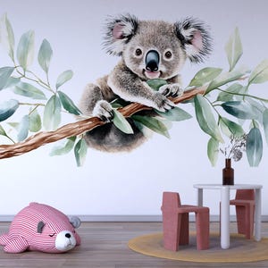 Peut inclure: Une peinture murale à l'aquarelle représentant un koala sur une branche avec des feuilles vertes. Un coussin en forme d'ours à rayures roses et blanches est posé sur le sol. Une petite table blanche et deux chaises roses complètent la scène.