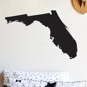 Puede incluir: Una silueta negra del estado de Florida se muestra en una pared blanca. La imagen es un diseño minimalista, perfecto para la decoración del hogar. Debajo, almohadas con patrones geométricos añaden un toque moderno.