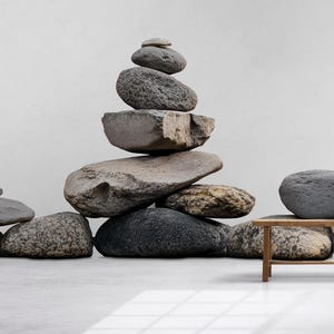 May include: A balanced stack of gray and brown stones, with a smaller stack to the left. A small wooden table holds a vase and a small stone. The background is a light wall and a wooden panel.