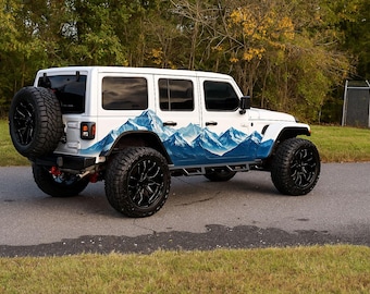 May include: A white SUV with black wheels and a spare tire. The vehicle features a mountain range graphic in blue and white along the side. The SUV is parked on a paved road with a grassy area in the foreground.