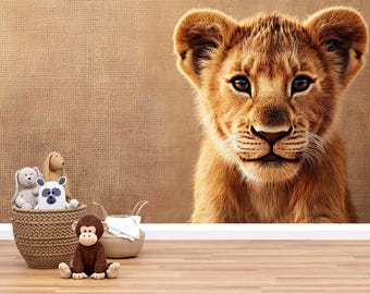 May include: A brown and tan lion cub with a soft, furry face looks directly at the camera. The cub is sitting on a brown textured background.