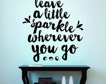 Può includere: Adesivo murale nero con la frase "leave a little sparkle wherever you go" in un carattere corsivo. L'adesivo è su una parete azzurra sopra un tavolo nero con oggetti decorativi.