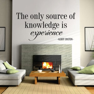 May include: A modern living room with two light gray sofas, each with green and gray pillows. A wooden coffee table sits in front of a fireplace with a lit fire. The wall above the fireplace has the quote "The only source of knowledge is experience" by Albert Einstein.