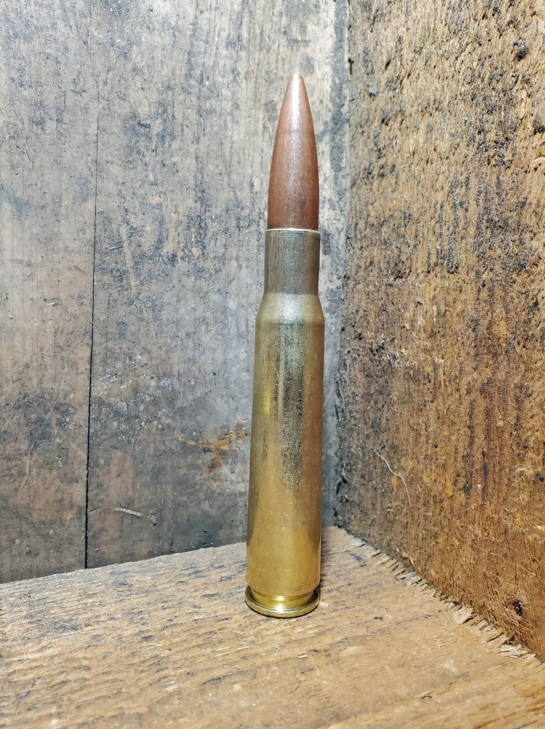 May include: A single, large bullet with a copper-colored tip and a brass casing. The bullet is standing upright against a wooden background. The casing is a shiny gold color.